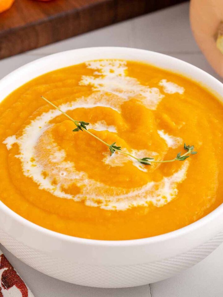 Close up of creamy butternut squash soup in a white bowl with a splash of coconut milk and thyme sprig on top
