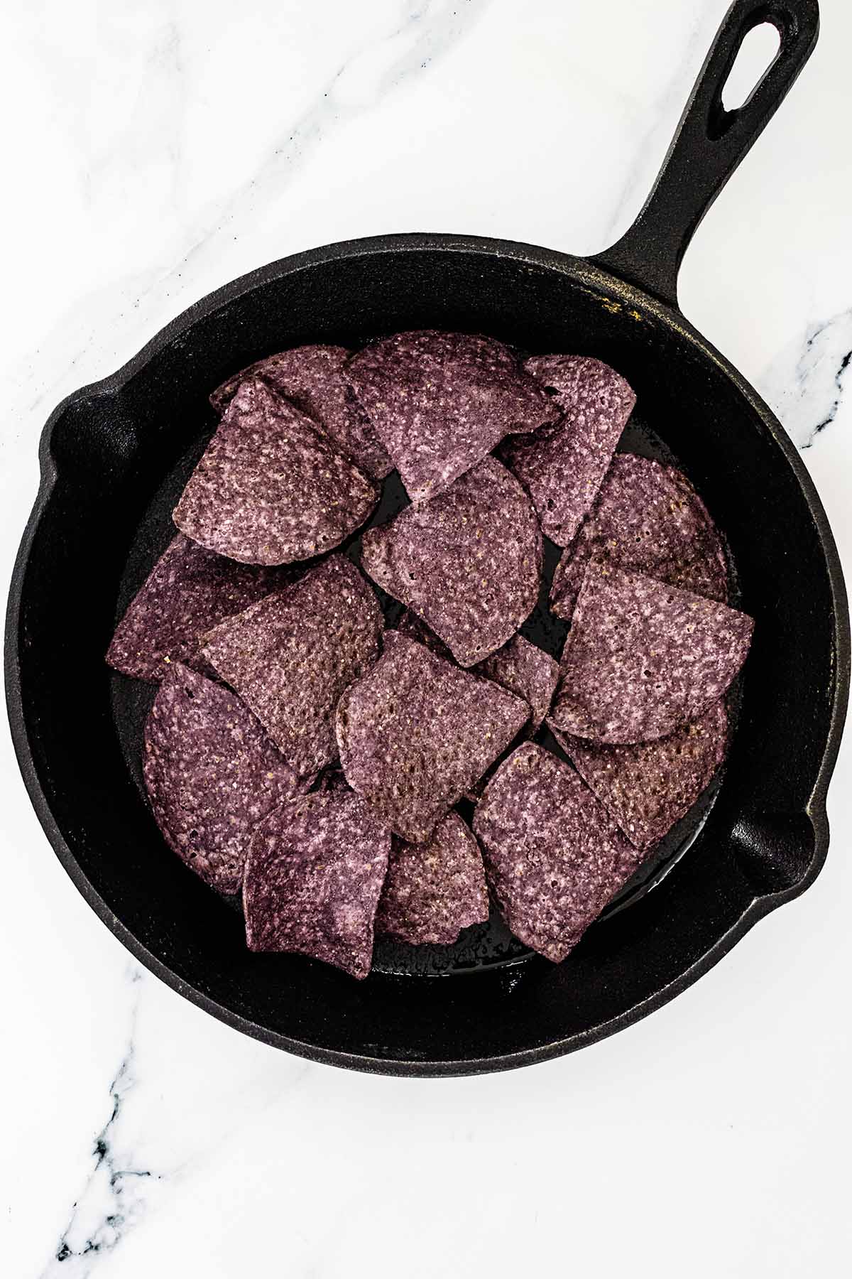 Overhead view of blue corn tortilla chips added to a cast iron skillet on a white marble background.