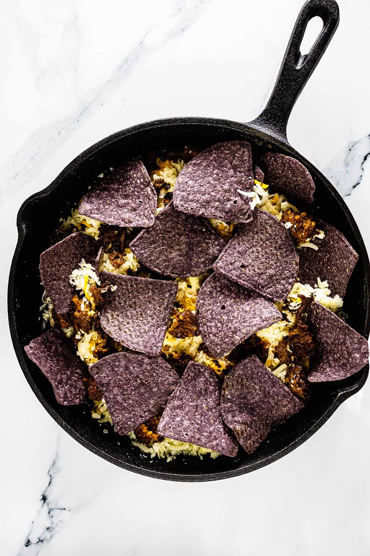 Overhead view of a layer of blue corn tortilla chips added to a skillet on a white marble background.