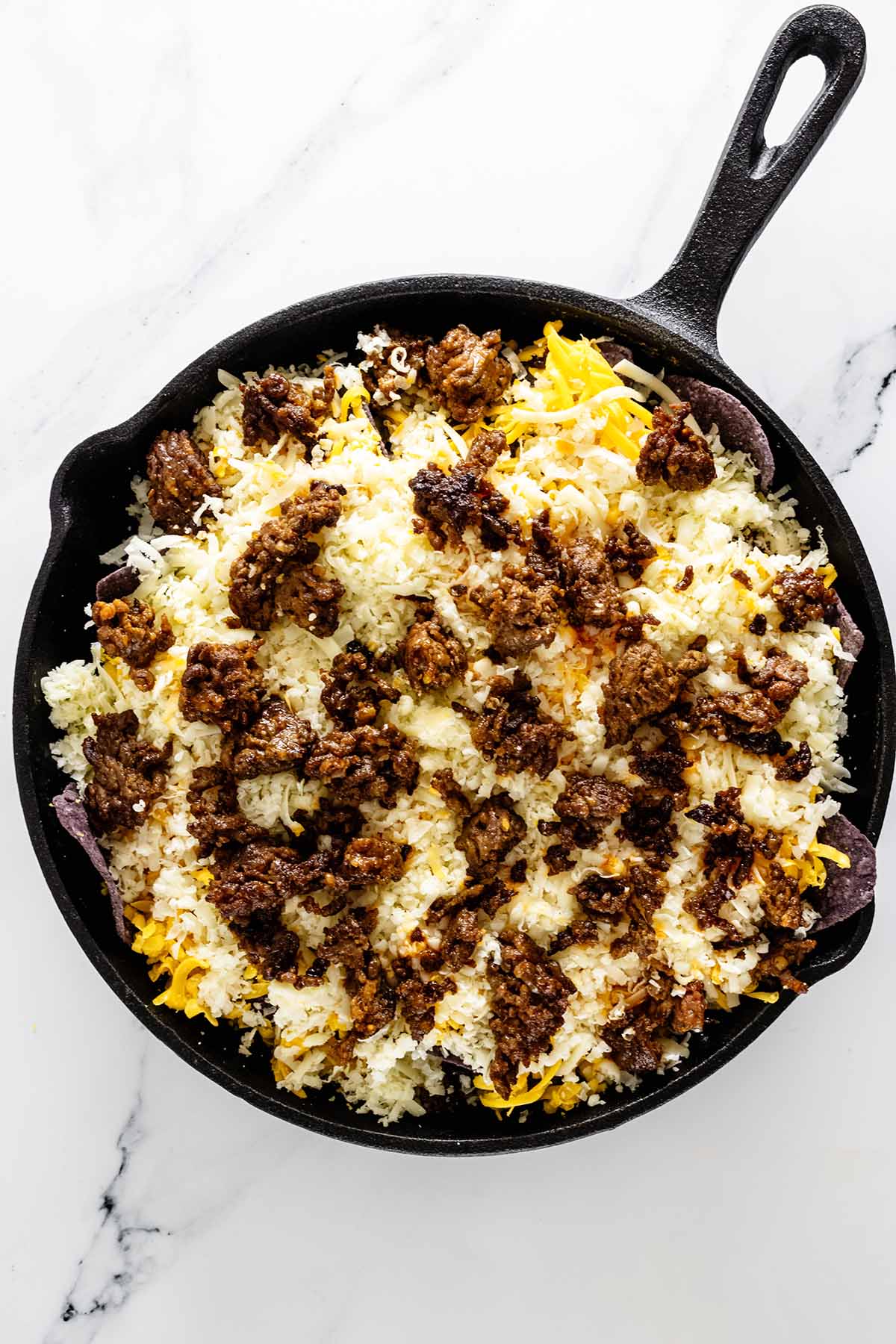 Overhead view of chorizo and shredded cheese added to a skillet on a white marble background.