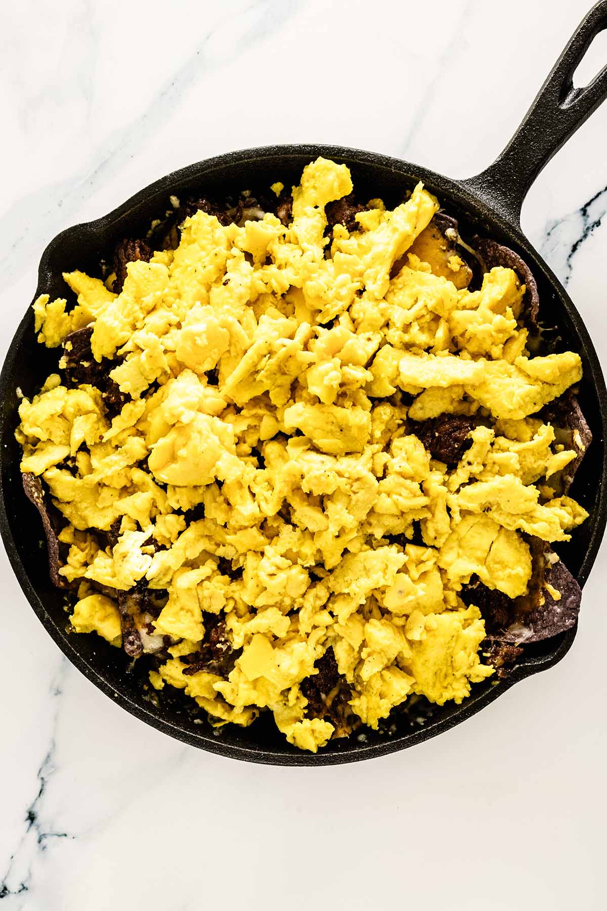 Overhead view of scrambled eggs added to cast iron skillet on white marble background.