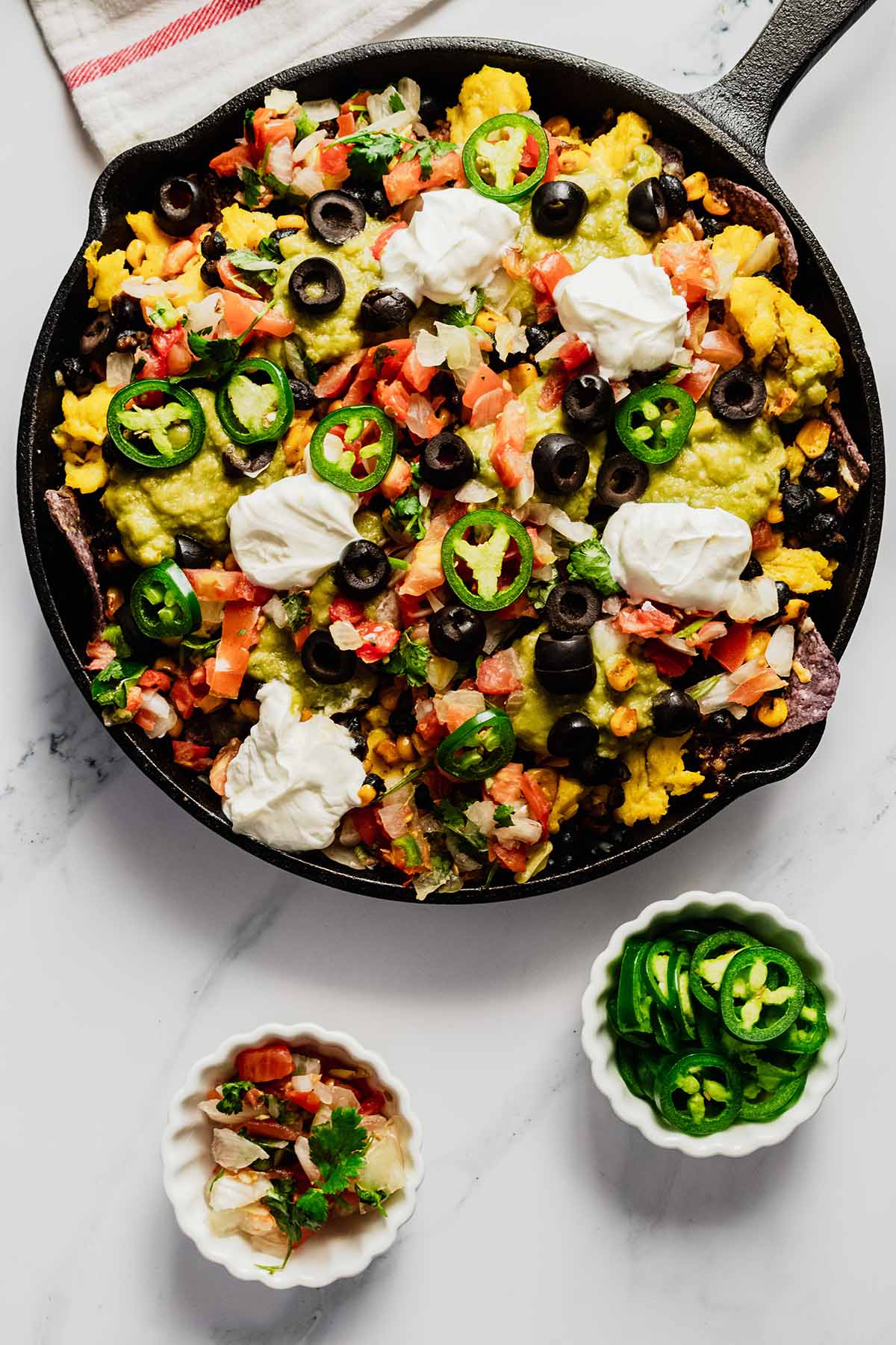 Overhead view of scrambled egg breakfast nachos in a cast iron skillet on a marble background with sliced jalapeños and pico de gallo in two small white bowls.