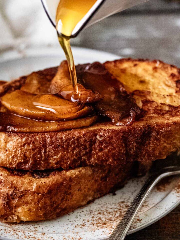 Maple syrup being poured over two slices of sourdough French toast topped with sautéed apples.