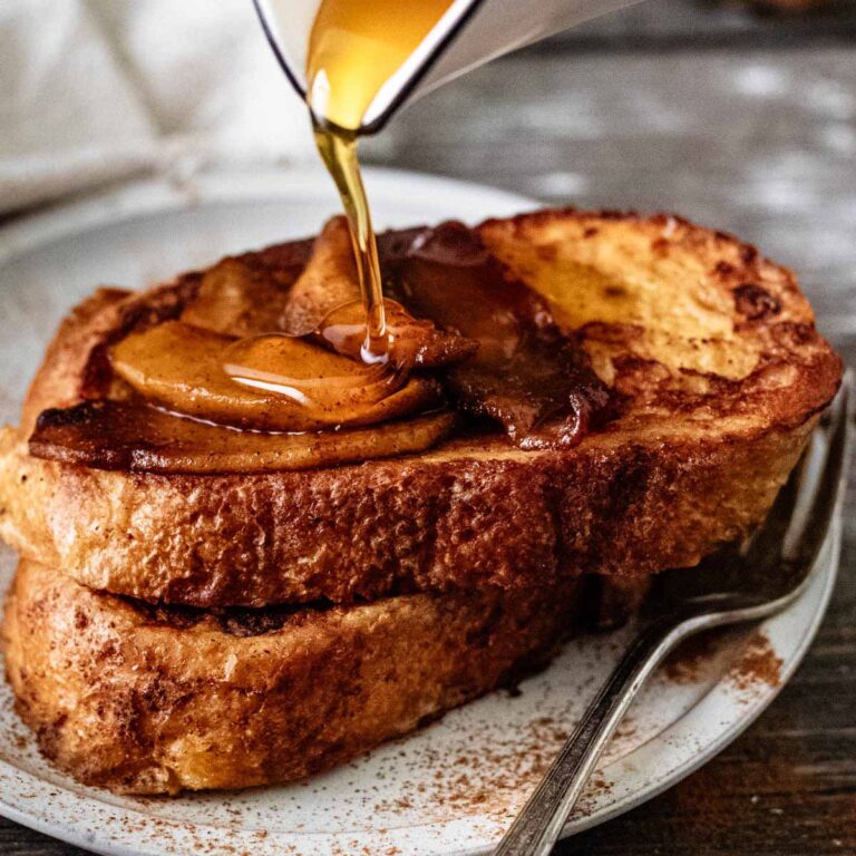 Maple syrup being poured over two slices of sourdough French toast topped with sautéed apples.