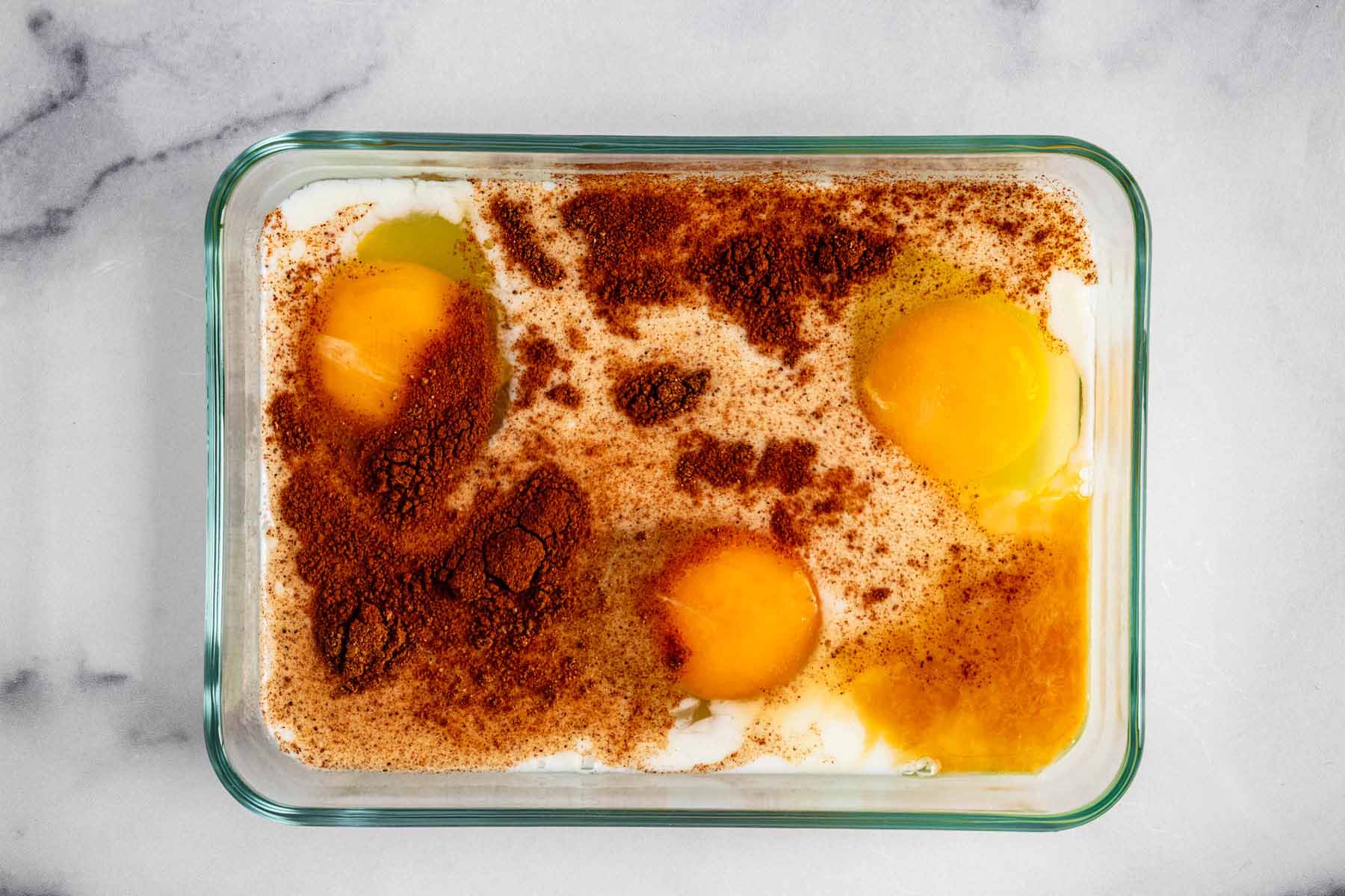 Eggs, milk, sugar, vanilla extract, cinnamon, and nutmeg in a small glass rectangular container.