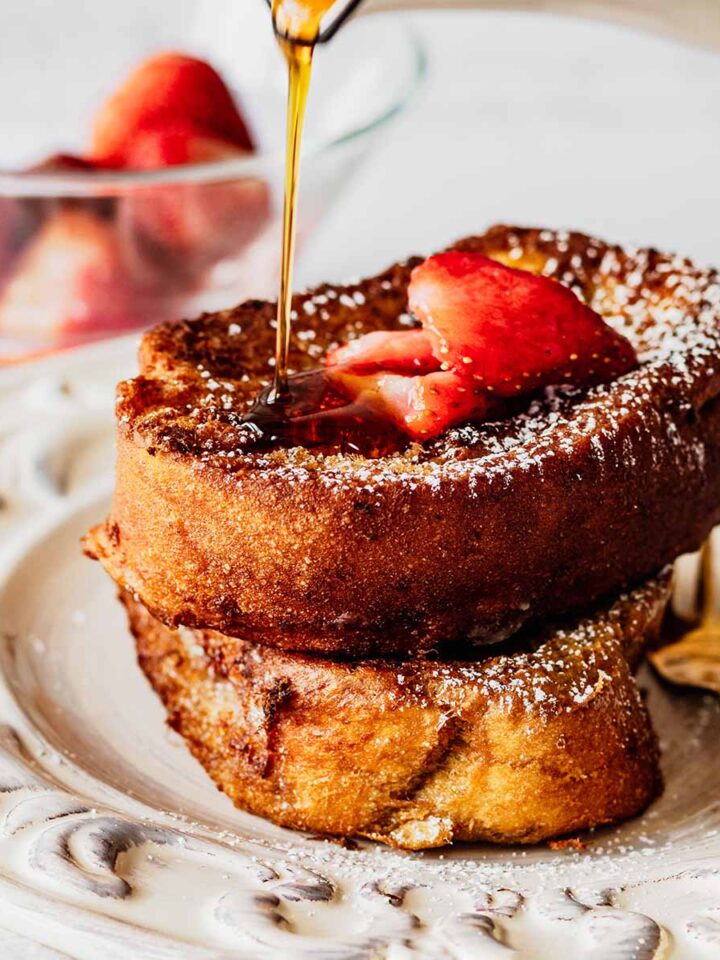 Syrup being poured on deep fried French toast topped with strawberries on a white plate with a gold fork.
