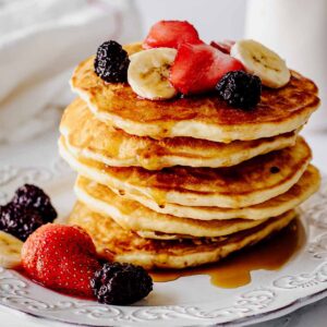 Stack of homemade pancakes topped with berries and sliced bananas on a white plate