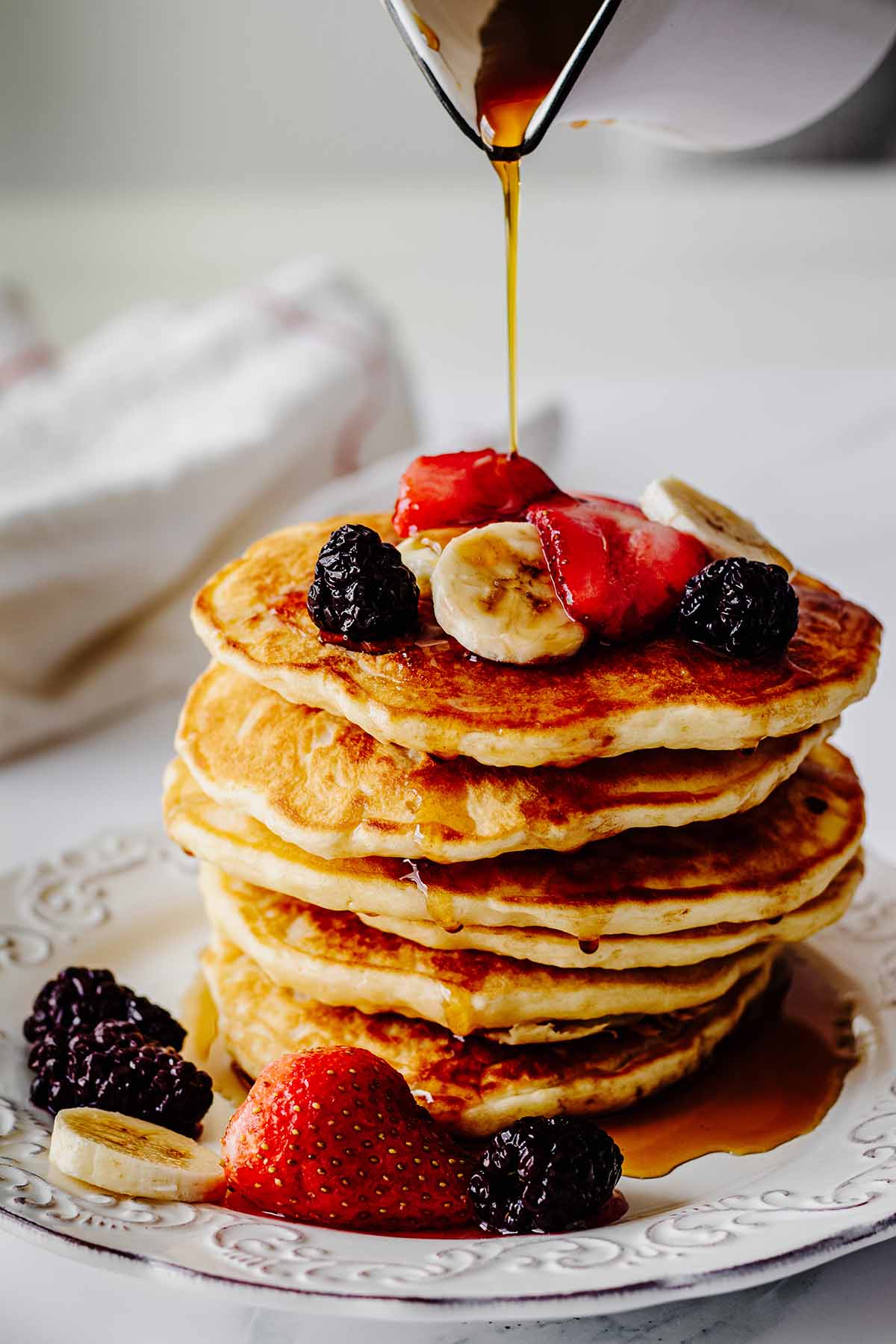 Syrup being poured on a stack of homemade pancakes topped with berries and sliced bananas on a white plate