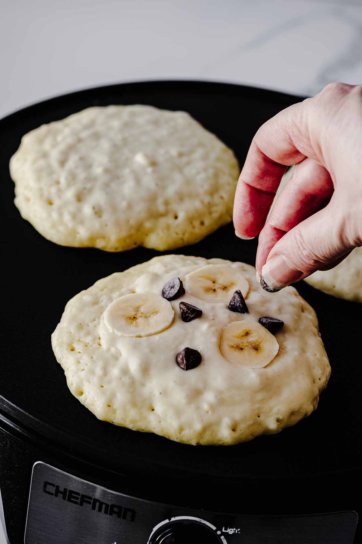 Chocolate chips and sliced bananas being placed on uncooked side of a pancake