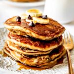 Stack of banana chocolate chip pancakes topped with sliced bananas, chocolate chips and maple syrup on a white plate with a gold fork