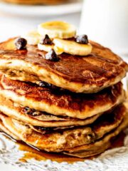 Stack of banana chocolate chip pancakes topped with sliced bananas, chocolate chips and maple syrup on a white plate with a gold fork