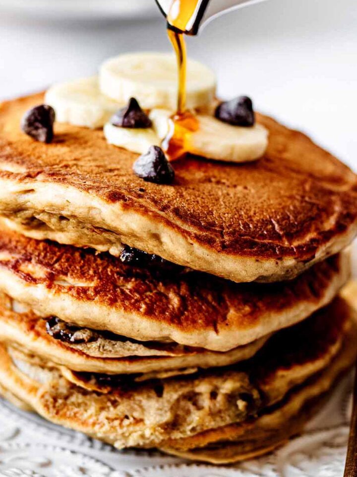 Maple syrup being poured on a stack of chocolate chip pancakes topped with sliced bananas and chocolate chips on a white plate