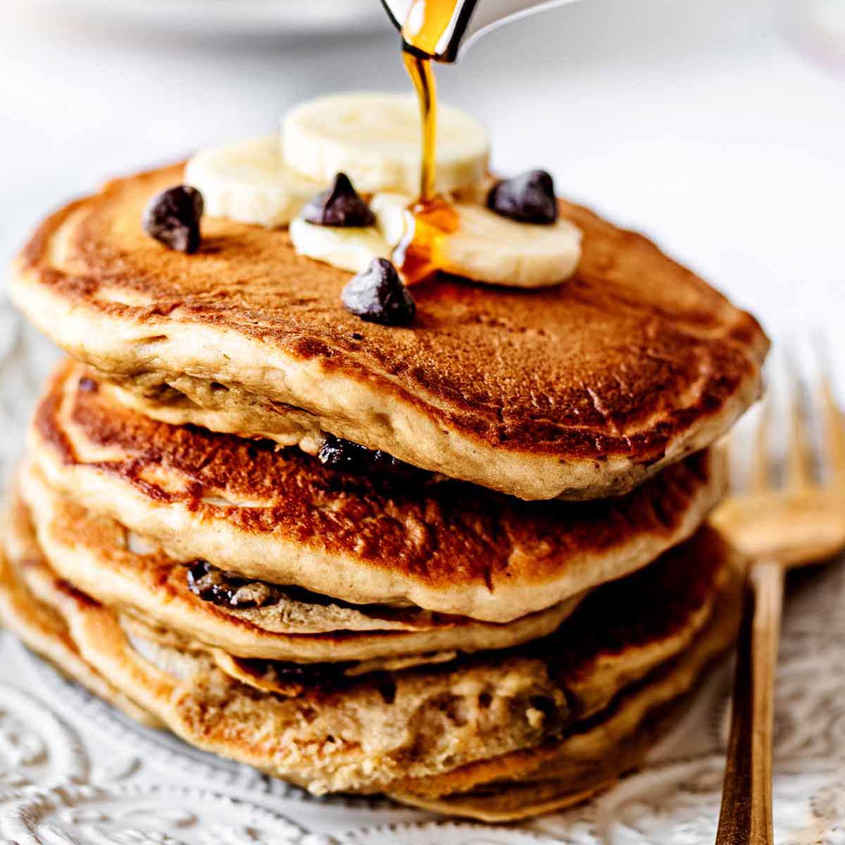 Maple syrup being poured on a stack of chocolate chip pancakes topped with sliced bananas and chocolate chips on a white plate