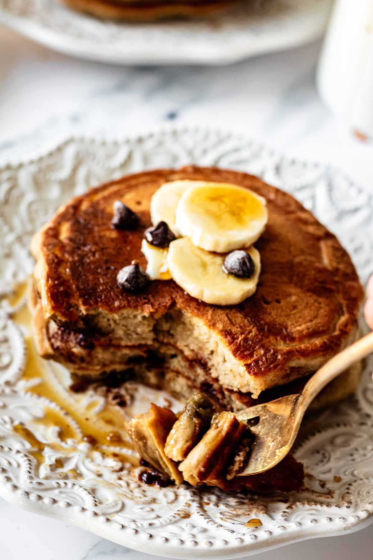 Short stack of banana pancakes on a white plate with a gold fork cutting a bite out