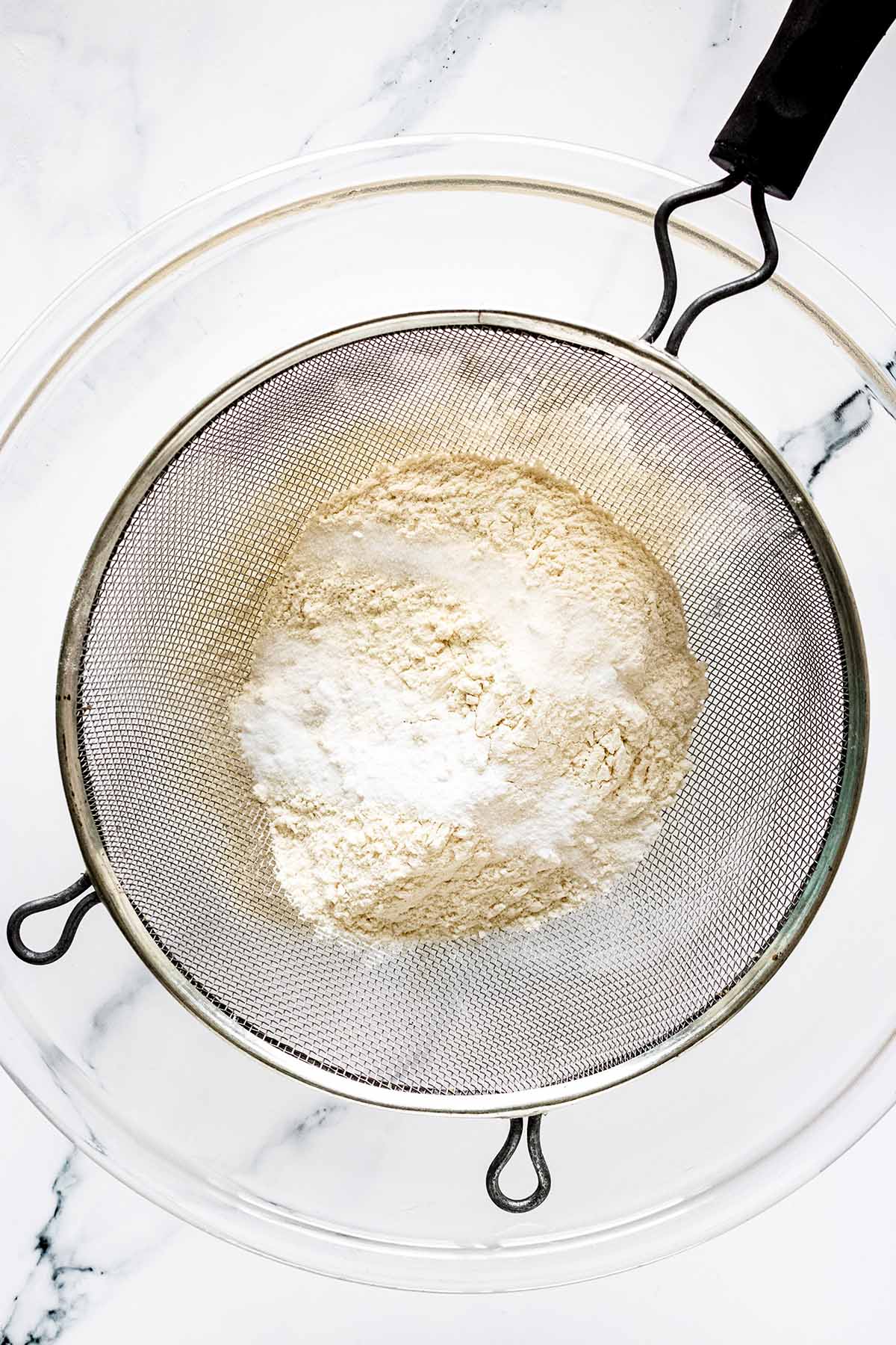 Overhead view of dry ingredients being sifted into a large glass bowl