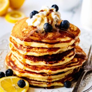 Stack of lemon blueberry pancakes on a white plate topped with whipped cream, maple syrup and blueberries