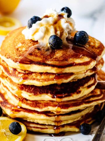 Stack of lemon blueberry pancakes on a white plate topped with whipped cream, maple syrup and blueberries
