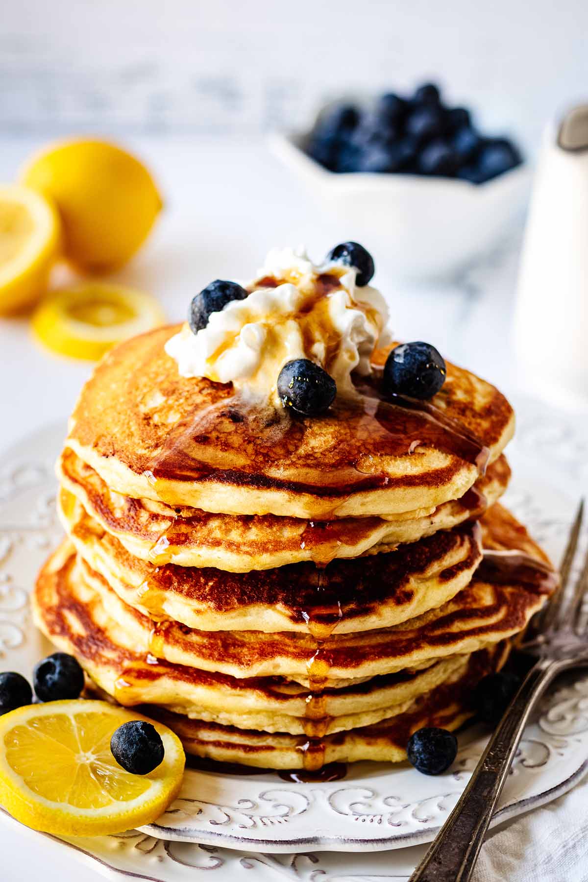 Stack of lemon blueberry pancakes on a white plate topped with whipped cream, maple syrup and blueberries