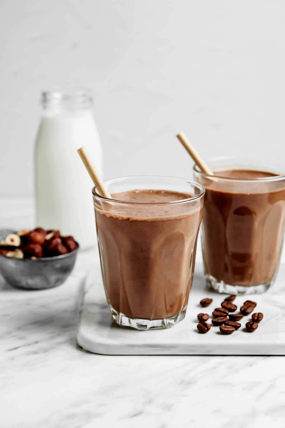 Chocolate protein shake in two glasses with straws and coffee beans on a white marble counter