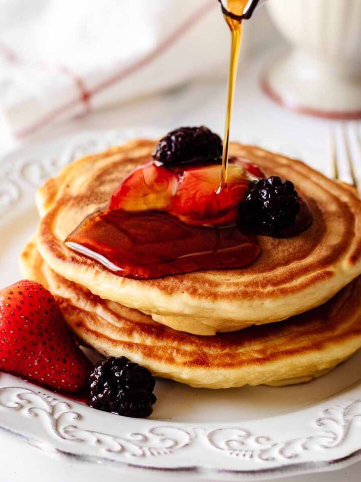 Syrup being poured on a small stack of pancakes topped with berries on a white plate
