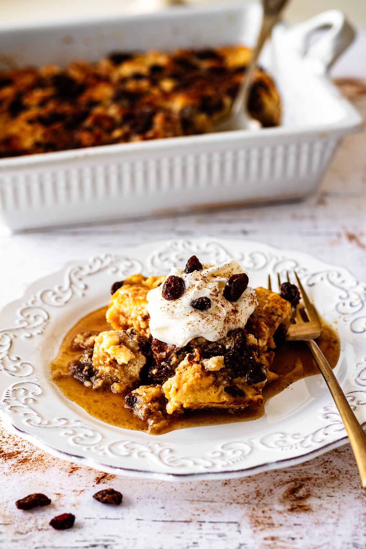 Serving of raisin bread pudding on a white plate topped with whipped cream and raisins