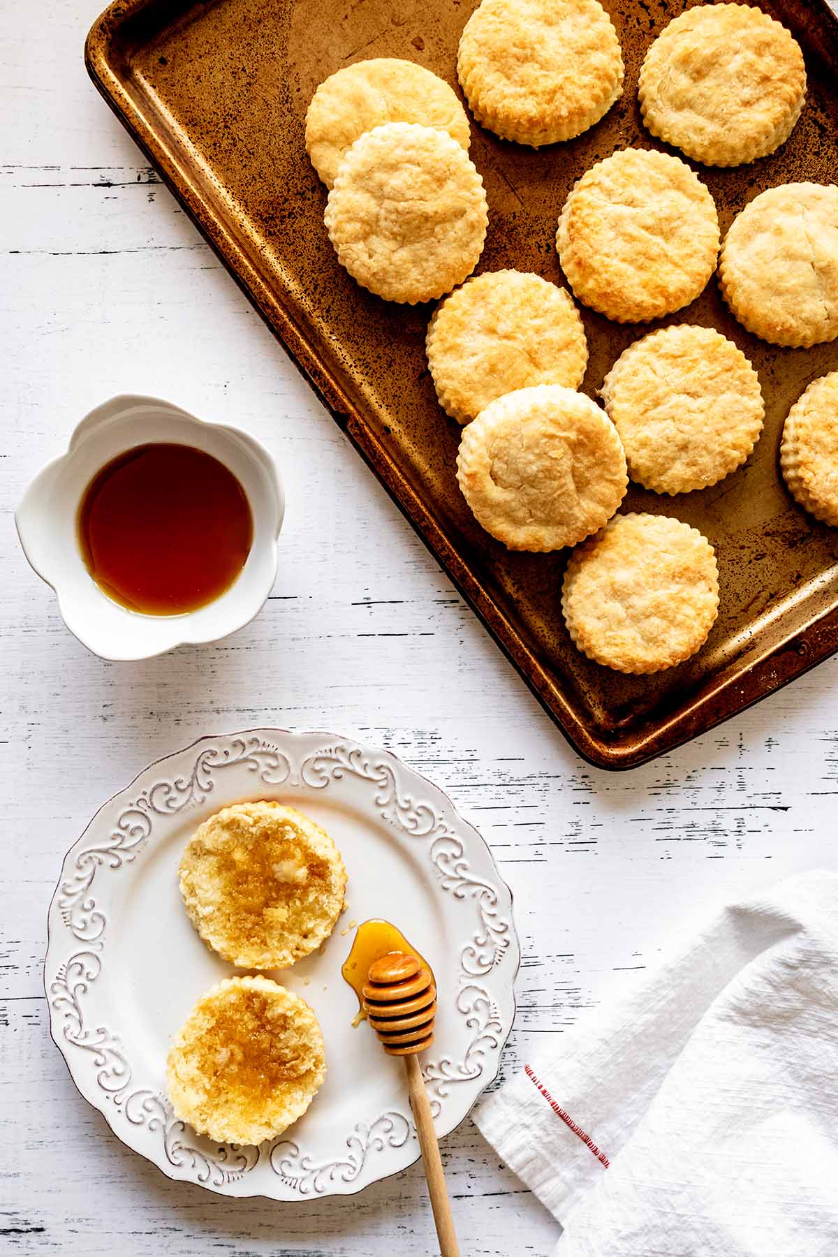 Overhead view of biscuits on a baking sheet and biscuits on a white plate with a bowl of honey