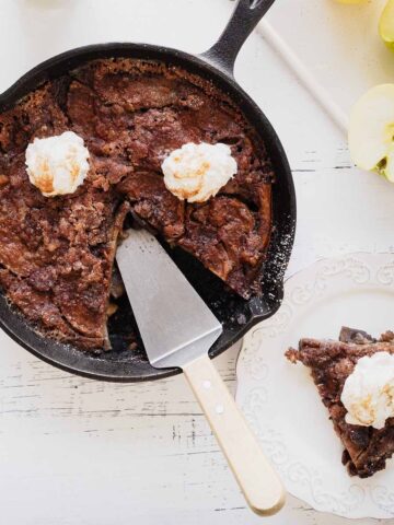 Overhead view of apple breakfast bake with a slice cut out in a cast iron skillet and a slice of whipped cream topped apple bake on a white plate