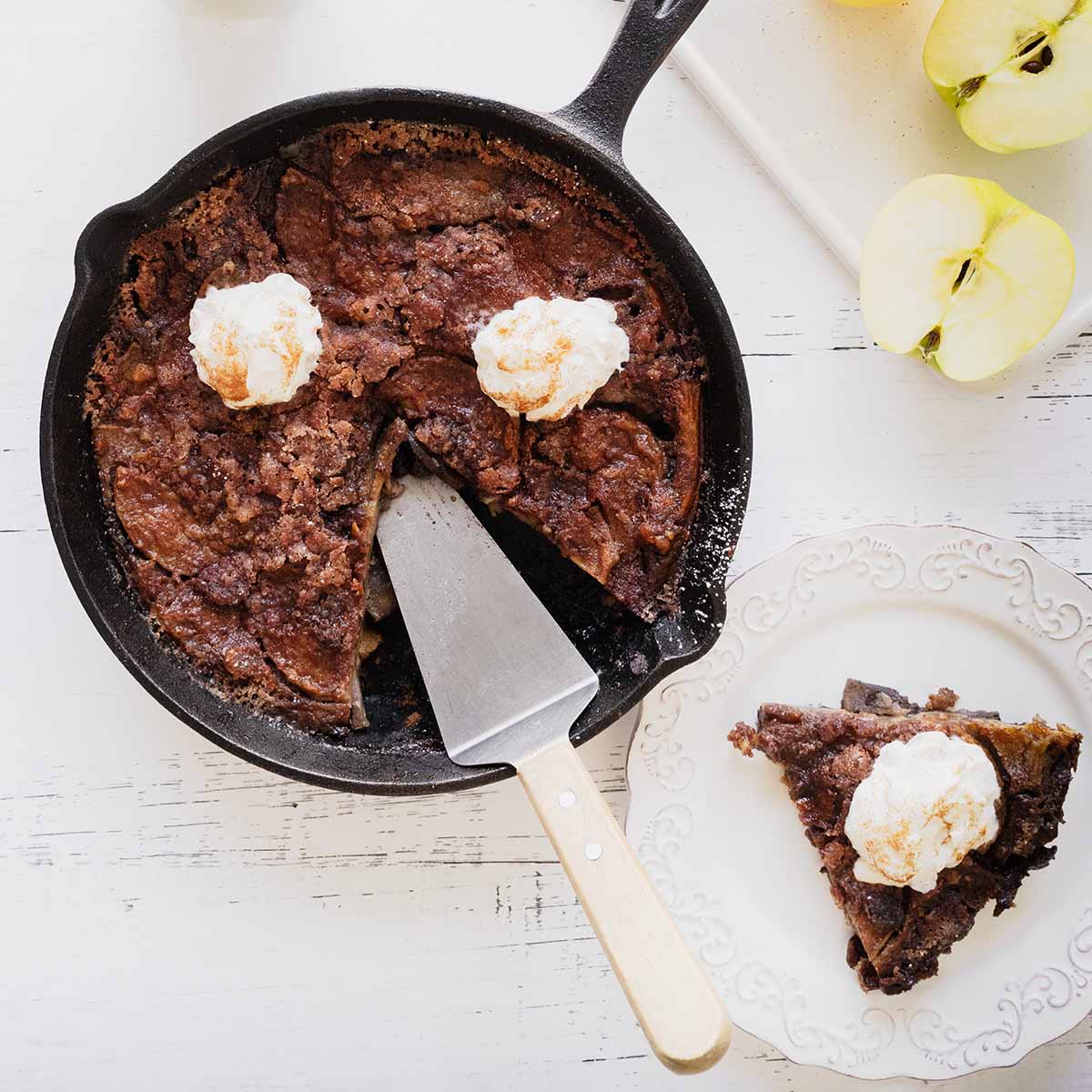 Overhead view of apple breakfast bake with a slice cut out in a cast iron skillet and a slice of whipped cream topped apple bake on a white plate