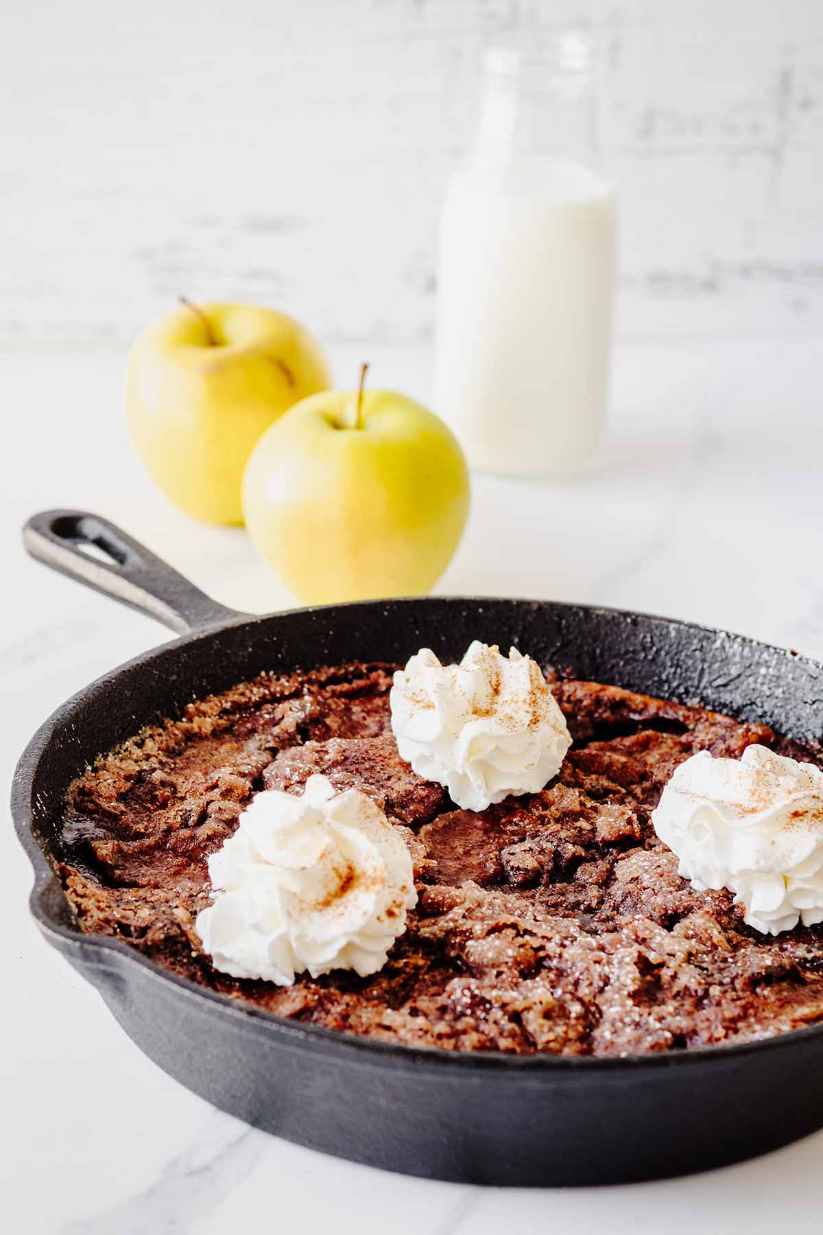 Apple breakfast bake topped with whipped cream in a cast iron skillet with apples and milk in the background