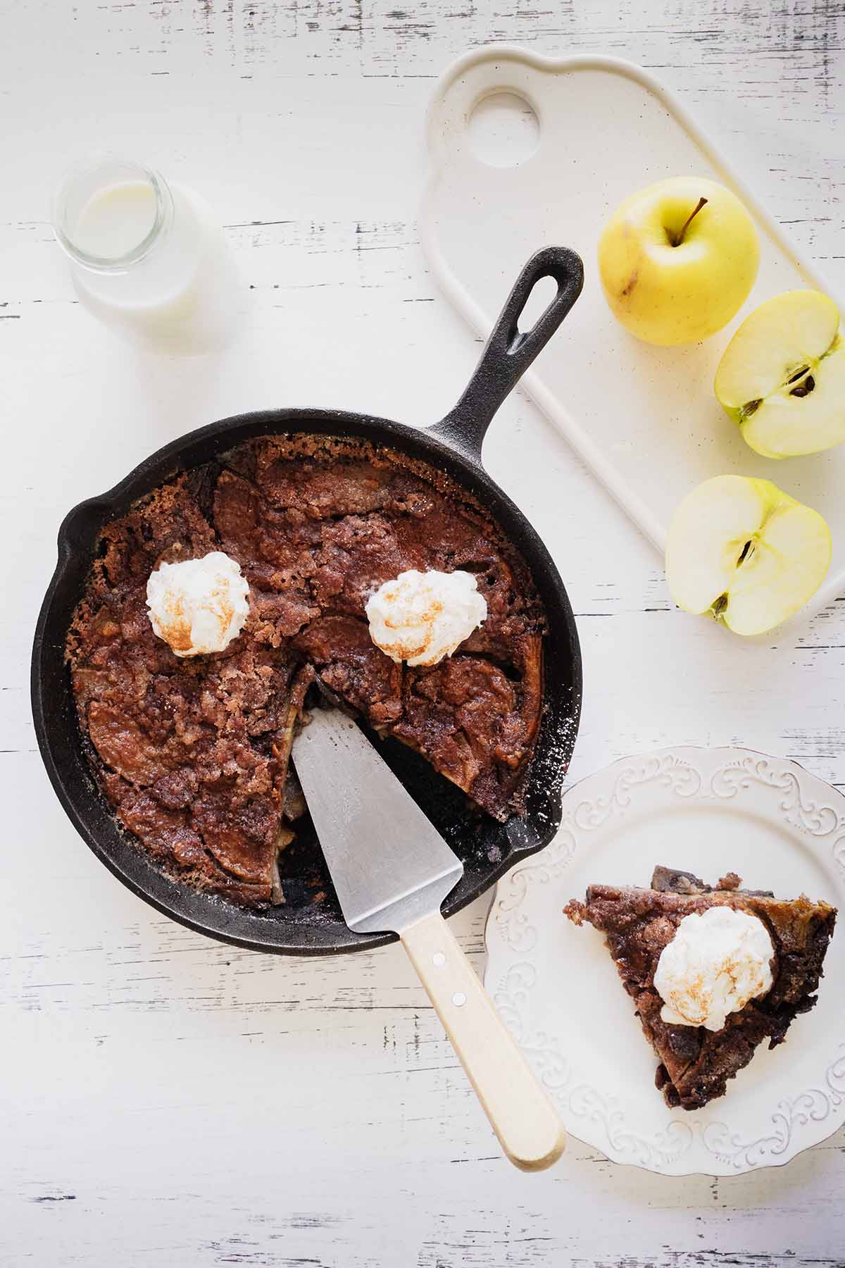 Overhead view of apple breakfast bake with a slice cut out in a cast iron skillet and a slice of whipped cream topped apple bake on a white plate