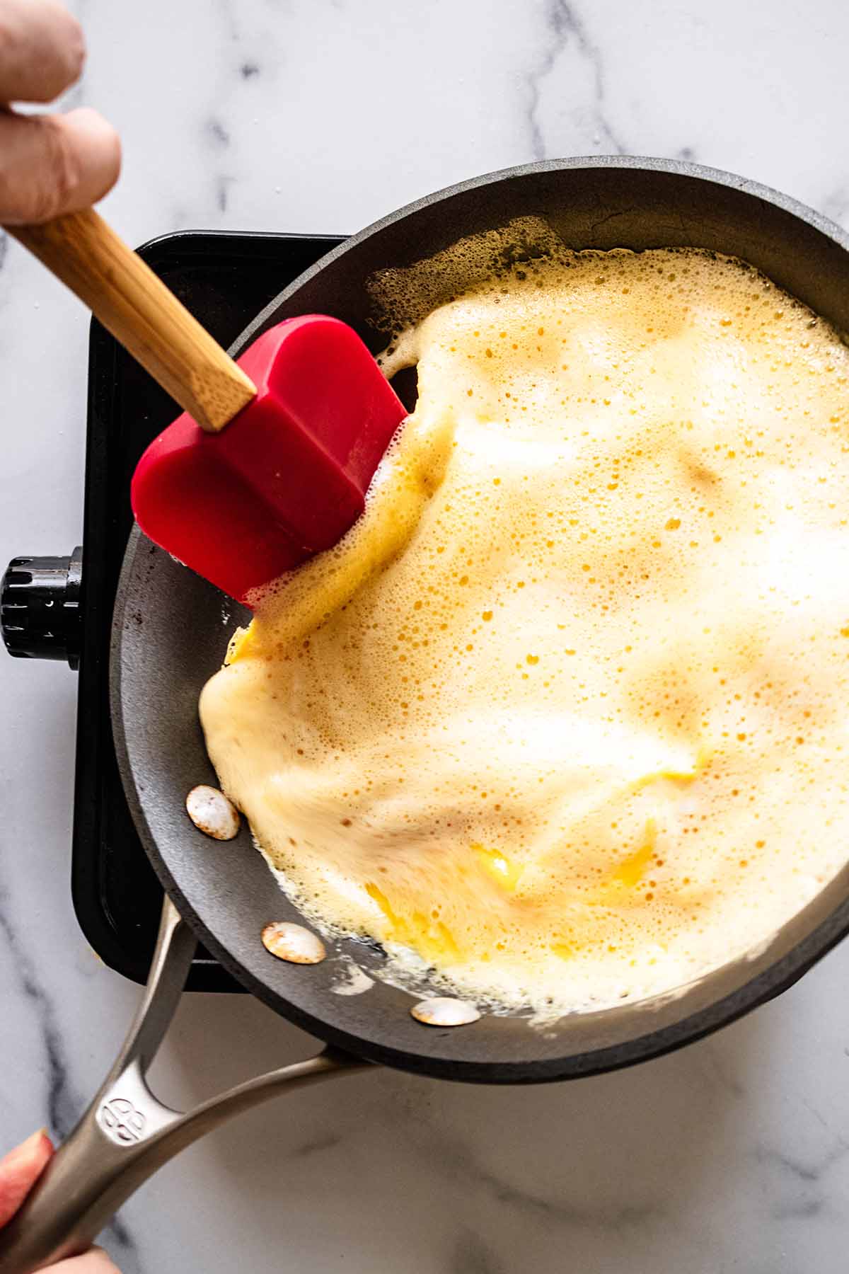 Red spatula lifting edges of partially-cooked omelette in a skillet. Skillet is being slightly tilted.