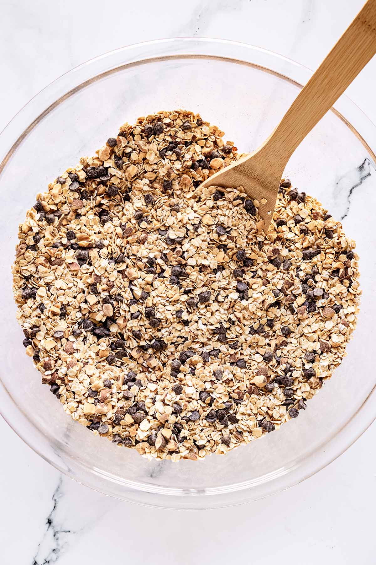 Overhead view of oat mixture in a glass bowl with a wooden spoon