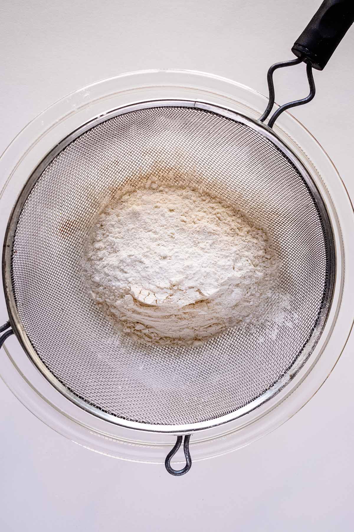 Overhead view of dry ingredients being sifted into a glass bowl