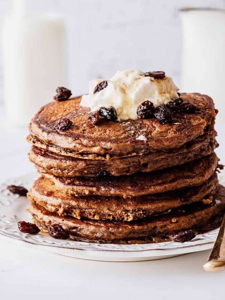 Stack of raisin pancakes topped with whipped cream and raisins on a white plate with a gold fork