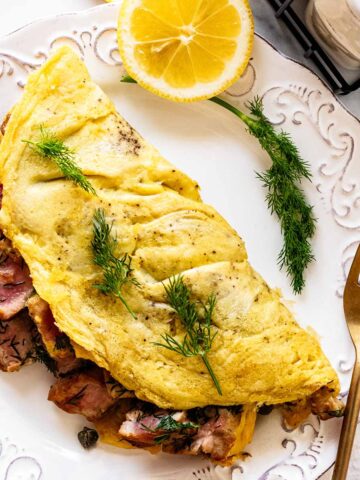 Overhead view of tuna omelette on a white plate with a sprig of dill and sliced lemons and a gold fork