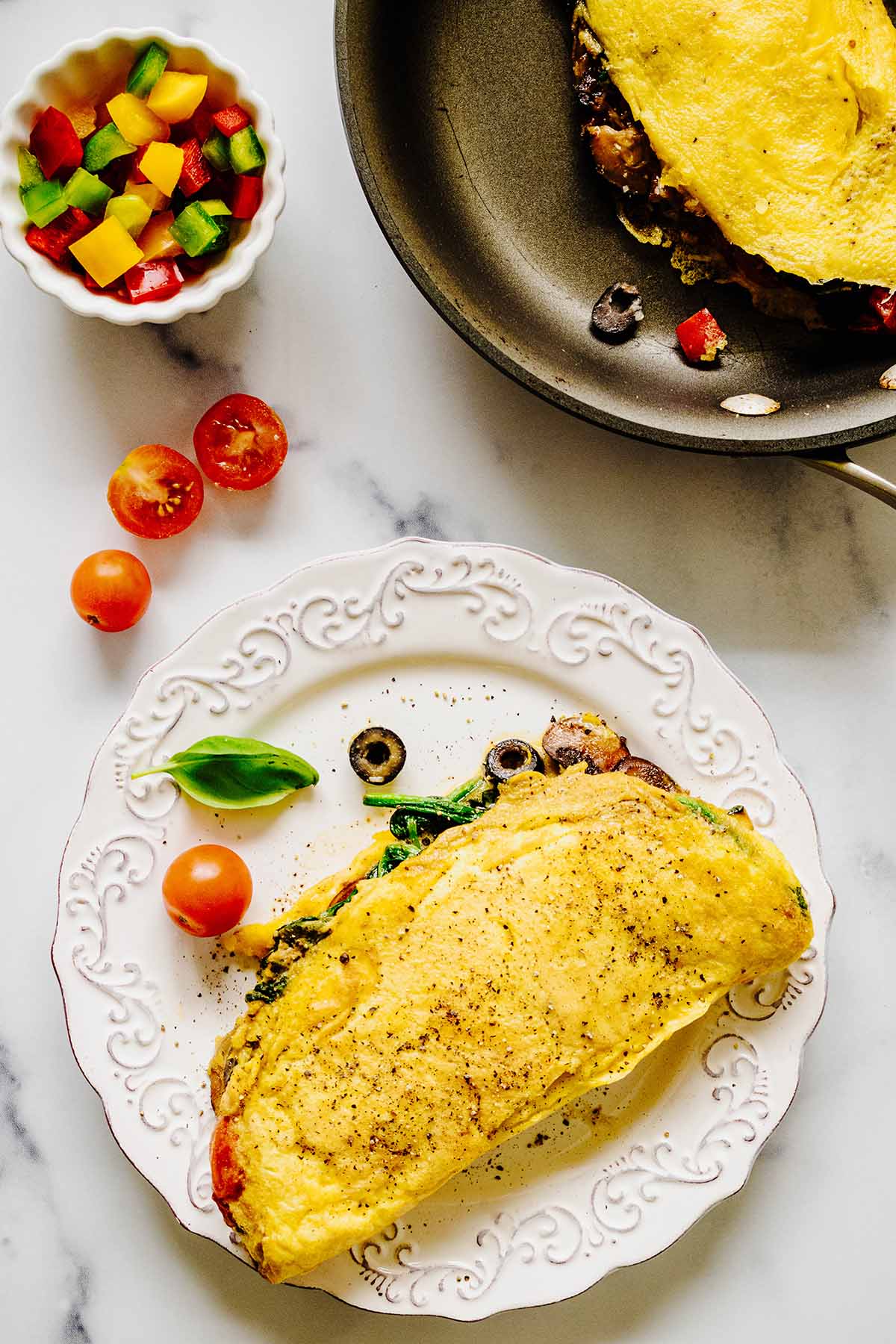 Overhead view of a vegetable omelette on a white plate with tomatoes and a small bowl of chopped peppers