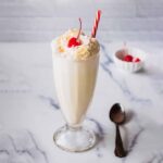 Coconut shake topped with whipped cream, toasted coconut, and a maraschino cherry in a tall glass with a red and white straw