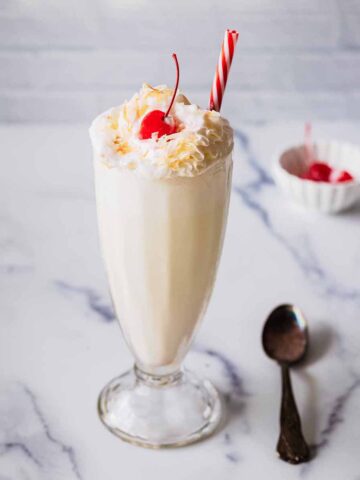 Coconut shake topped with whipped cream, toasted coconut, and a maraschino cherry in a tall glass with a red and white straw