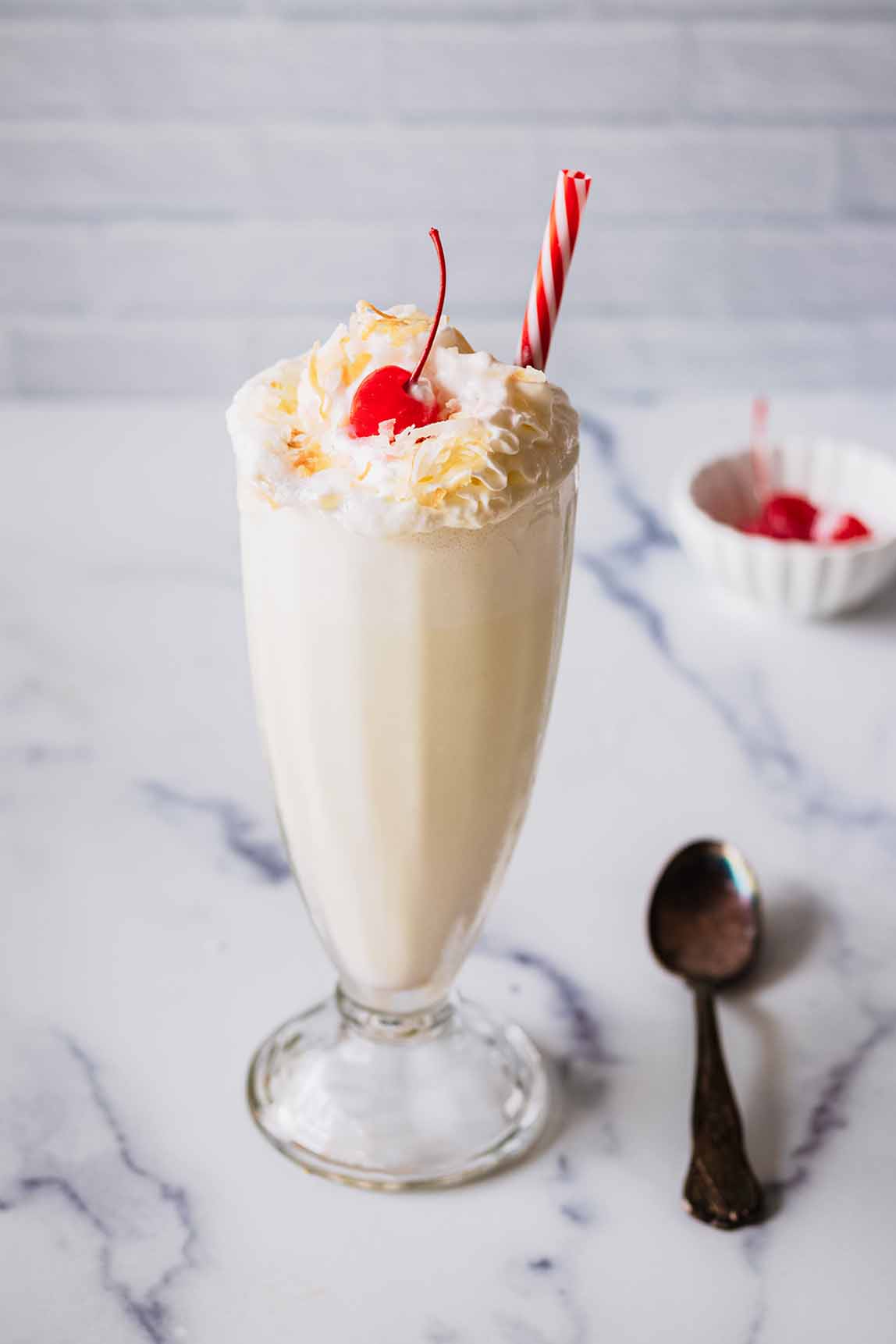 Coconut shake topped with whipped cream, toasted coconut, and a maraschino cherry in a tall glass with a red and white straw