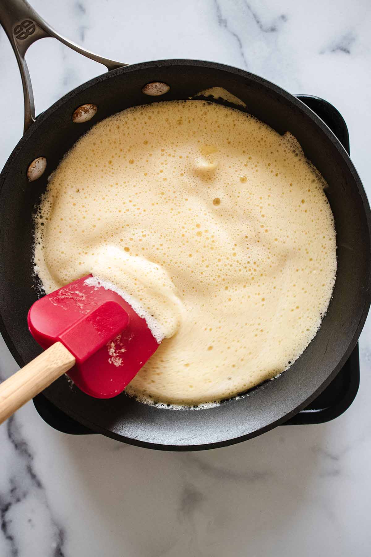 Overhead view of a red spatula gently pushing partially-cooked eggs toward the center of a skillet