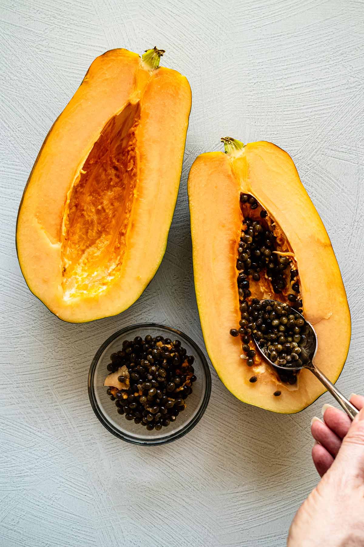 Overhead view of halved papaya on a countertop. A spoon is scooping out the seeds into a small glass bowl.