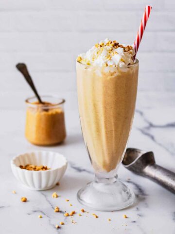 Peanut butter milkshake in a tall glass with whipped cream and a red and white straw.