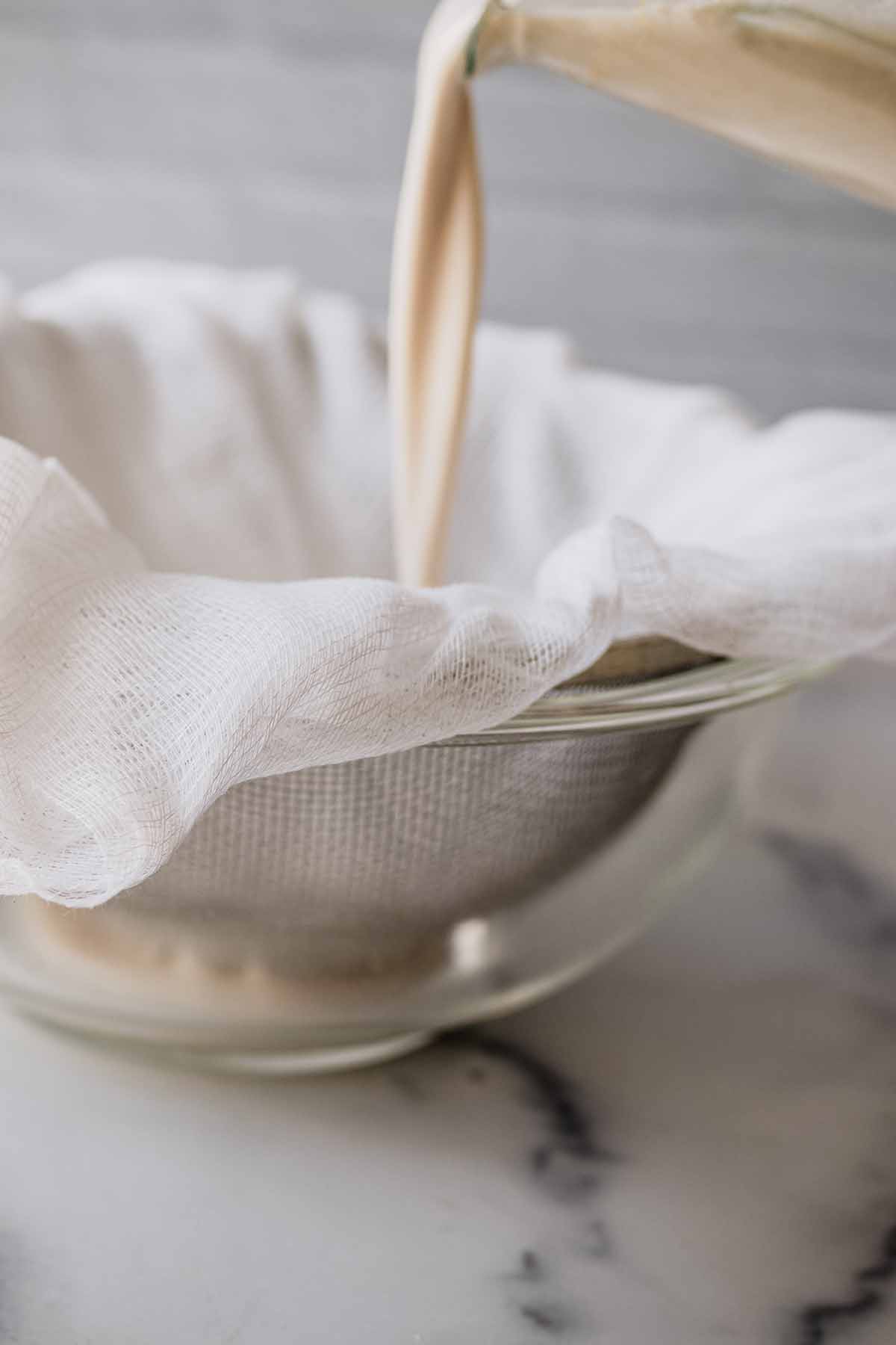 Nut milk being poured into a cheese cloth and mesh strainer into a glass bowl