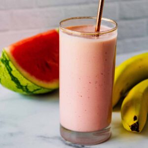 Smoothie in a tall glass with a bronze straw and watermelon slice and bananas in the background