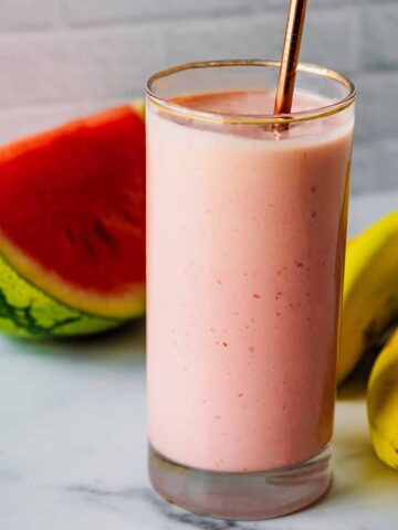 Smoothie in a tall glass with a bronze straw and watermelon slice and bananas in the background