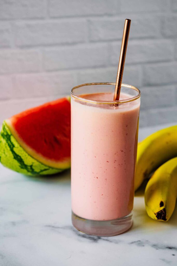 Watermelon banana smoothie in a tall glass with a bronze straw and watermelon slice and bananas in the background