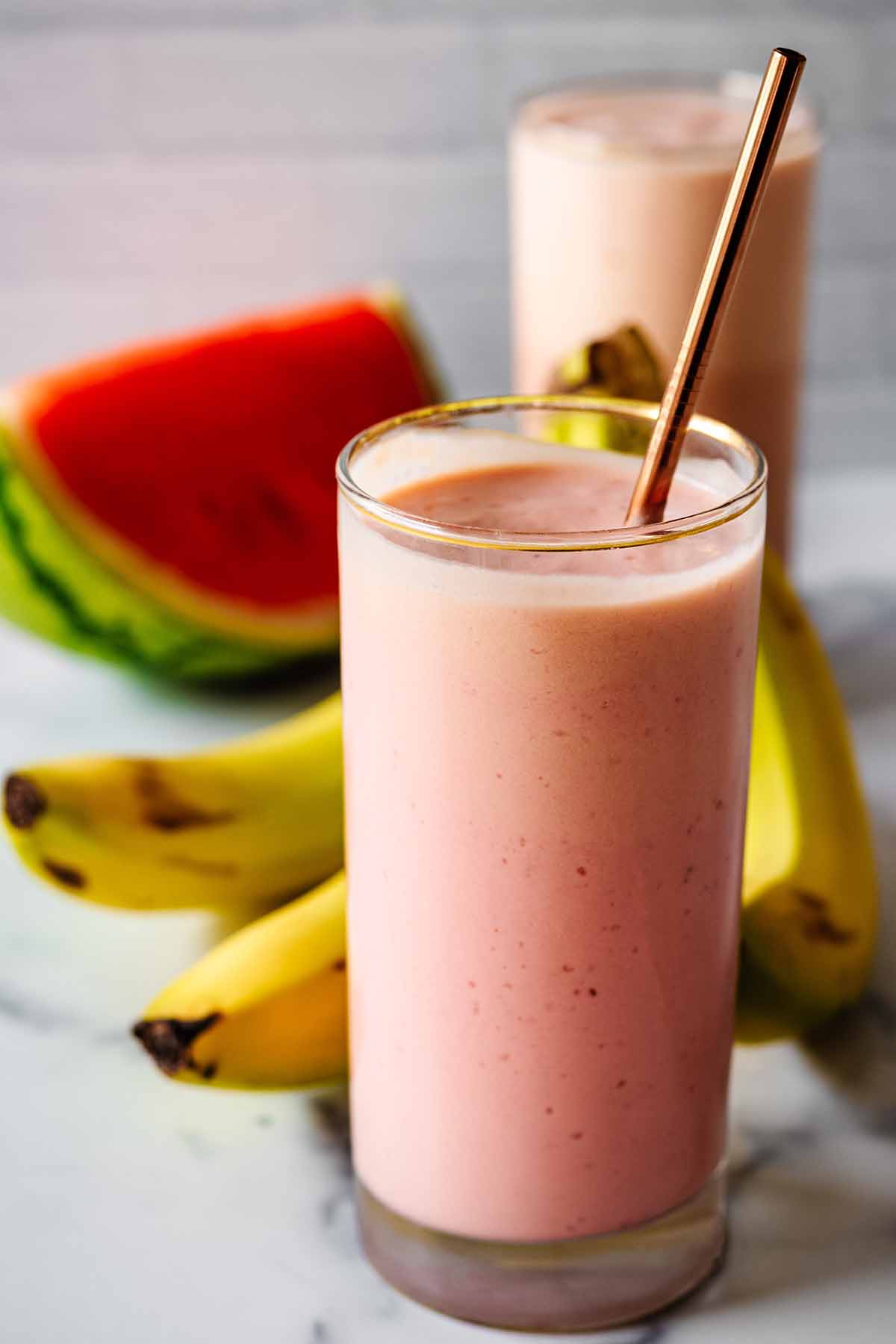 Watermelon banana smoothie in a tall glass with a bronze straw and a second smoothie, watermelon slice and bananas in the background