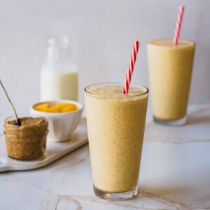 Two smoothies in tall glasses with red and white straws. A small jar of nut butter and small white bowl of chopped mango are off to the side.