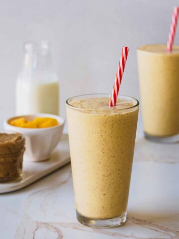 Two smoothies in tall glasses with red and white straws. A small jar of nut butter and small white bowl of chopped mango are off to the side.