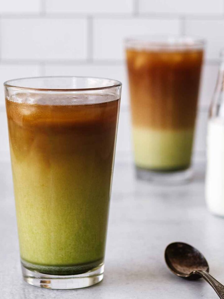 Two iced matcha coffee drinks in tall glasses with a small bottle of milk and a spoon